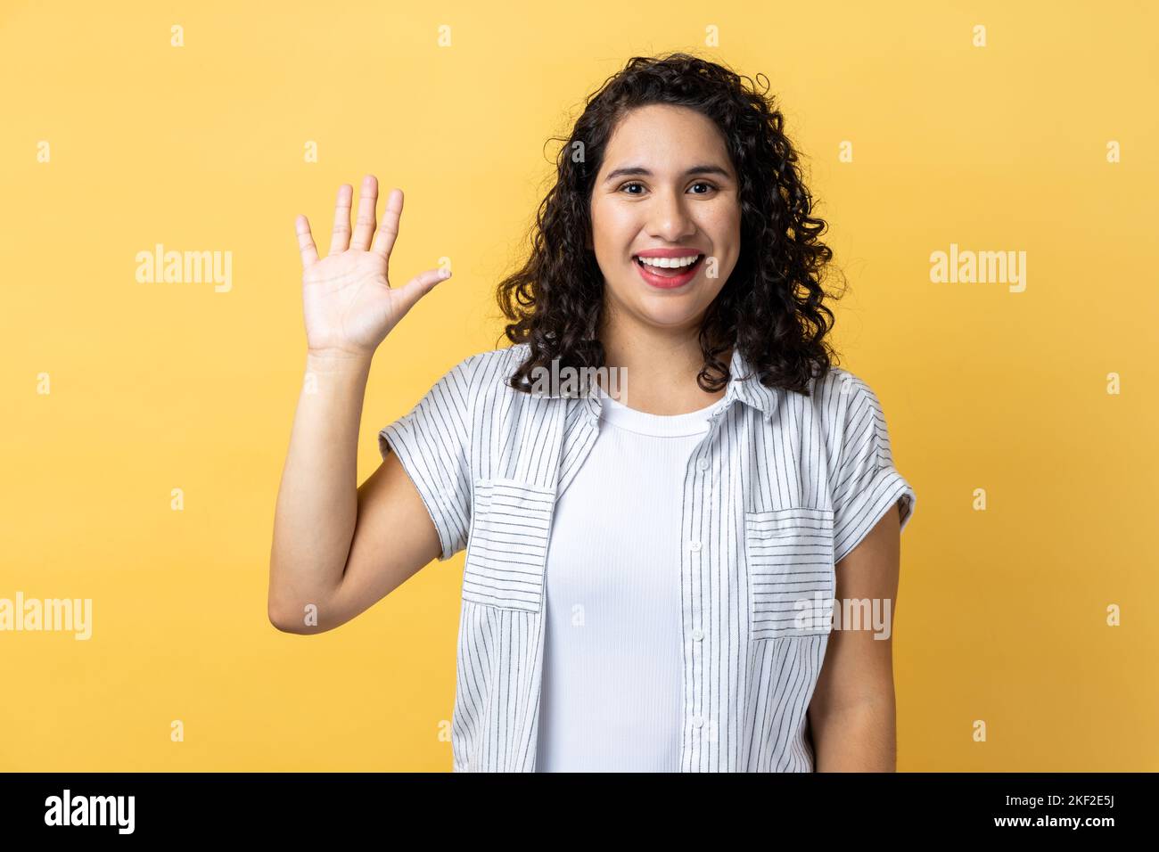 Portrait de sympathique positive belle femme avec les cheveux ondulés foncés sourit édeniement, soulève la paume salue ami, en disant bonjour ou Au revoir. Studio d'intérieur isolé sur fond jaune. Banque D'Images