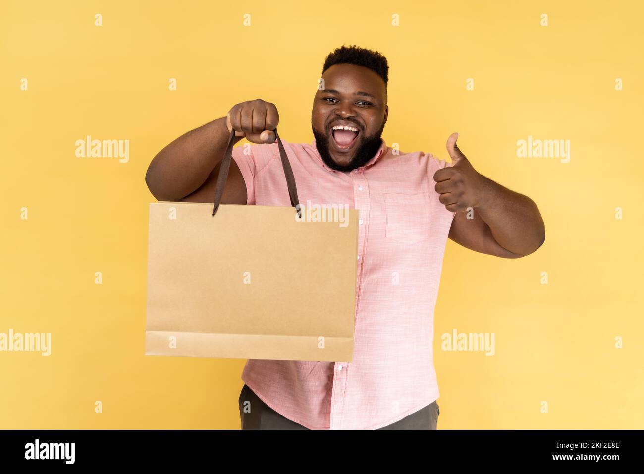 Portrait de l'homme portant une chemise rose montrant le pouce vers le haut tenant le sac de shopping, heureux avec de bons achats et des réductions, recommander. Studio d'intérieur isolé sur fond jaune. Banque D'Images