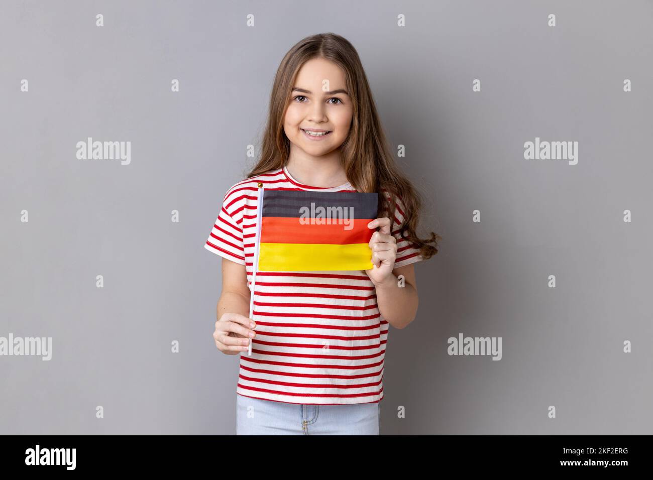 Drapeau de l'Allemagne. Portrait de satisfait optimiste adorable adorable adorable adorable petite fille portant un T-shirt rayé tenant le drapeau allemand, regardant l'appareil photo. Prise de vue en studio isolée sur fond gris. Banque D'Images