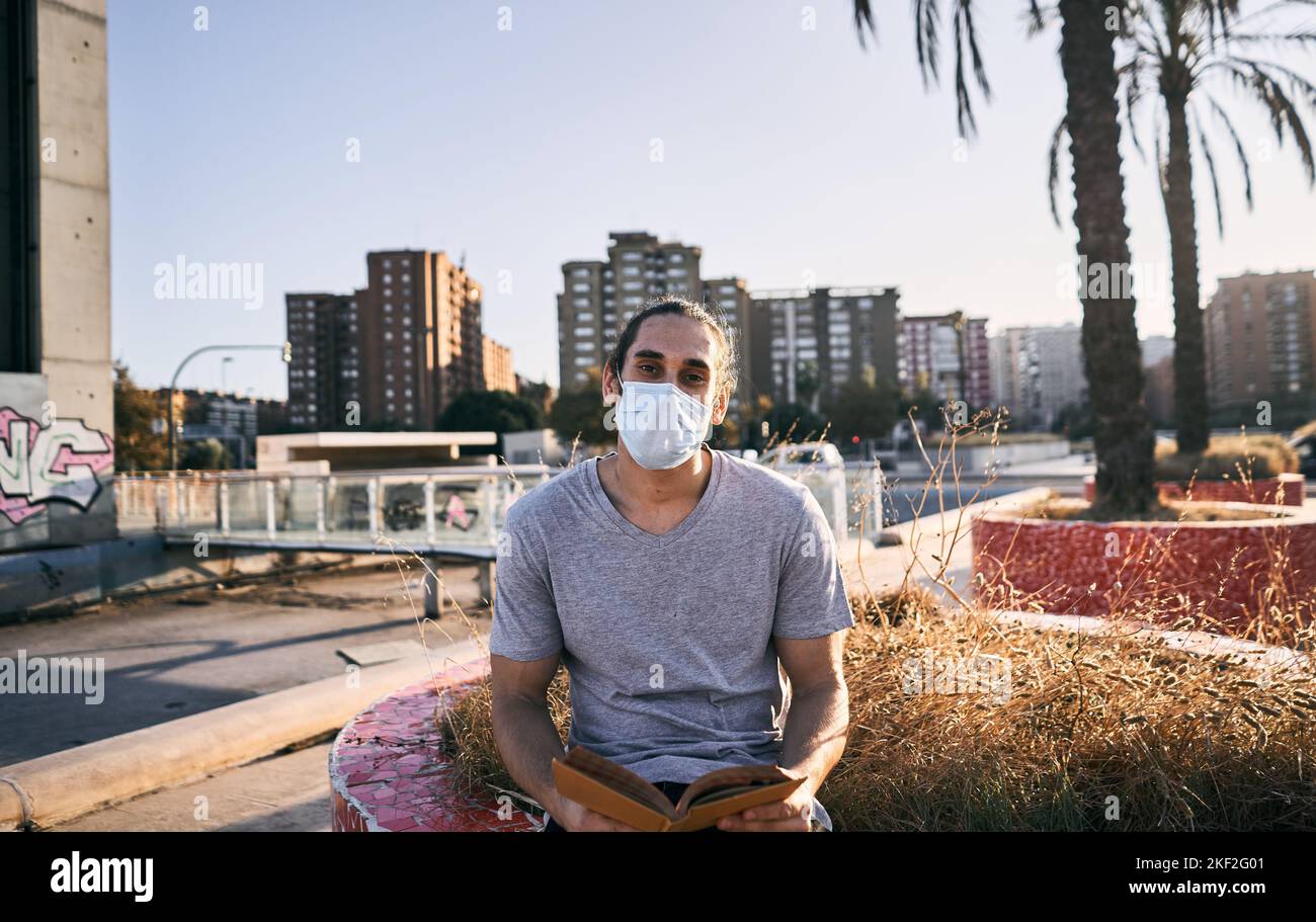 Jeune homme caucasien avec de longs cheveux recueillis en t-shirt gris masque de visage assis dans le parc regardant la caméra avec un livre dans ses mains dans la ville Banque D'Images