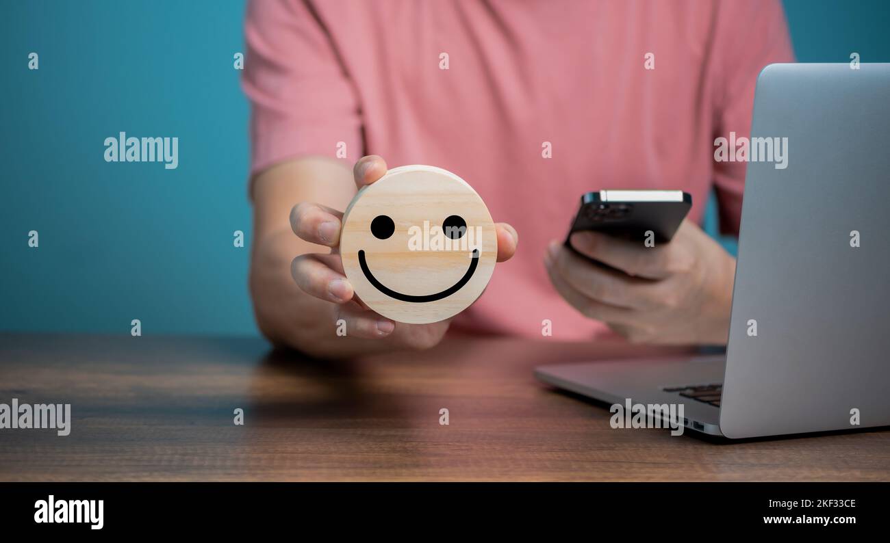 Homme utilisant un téléphone mobile et tenant un disque en bois avec des icônes de visage souriant, le service de satisfaction de la clientèle, les commentaires, la notation et l'avis positif de la clientèle, Banque D'Images