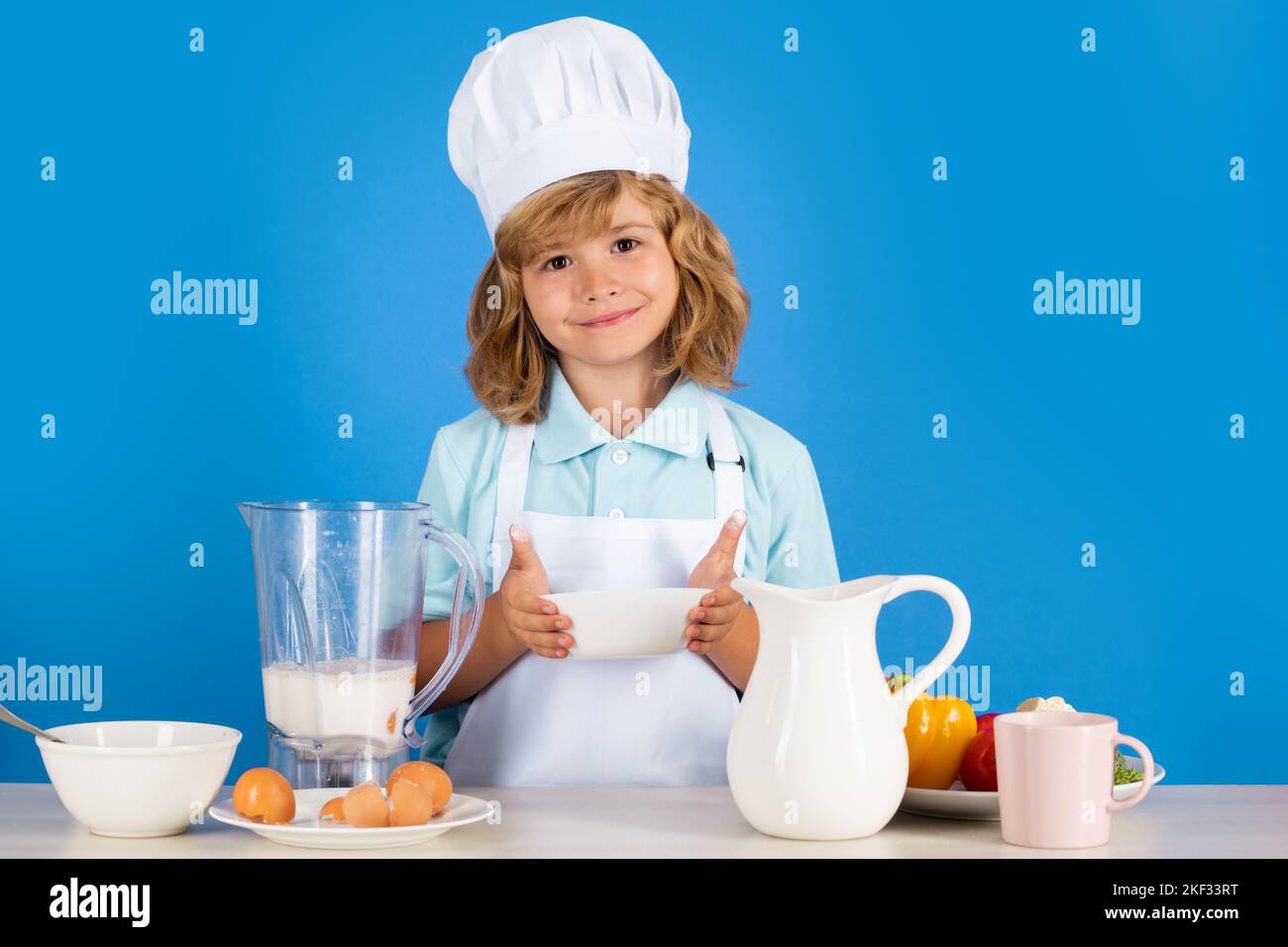 Portrait d'un enfant de 7 ans et 8 ans en chapeau et tablier de cuisine pour faire de la salade de fruits et cuisiner des aliments dans la cuisine. Mignon petit blond joyeux chef souriant. Banque D'Images