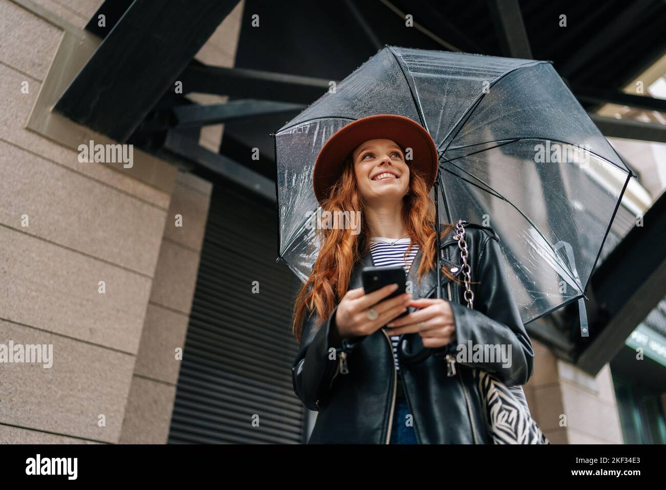 Vue en contre-angle de positive gingembre jeune femme portant un chapeau élégant en dactylographiant smartphone en regardant loin debout avec parapluie transparent Banque D'Images