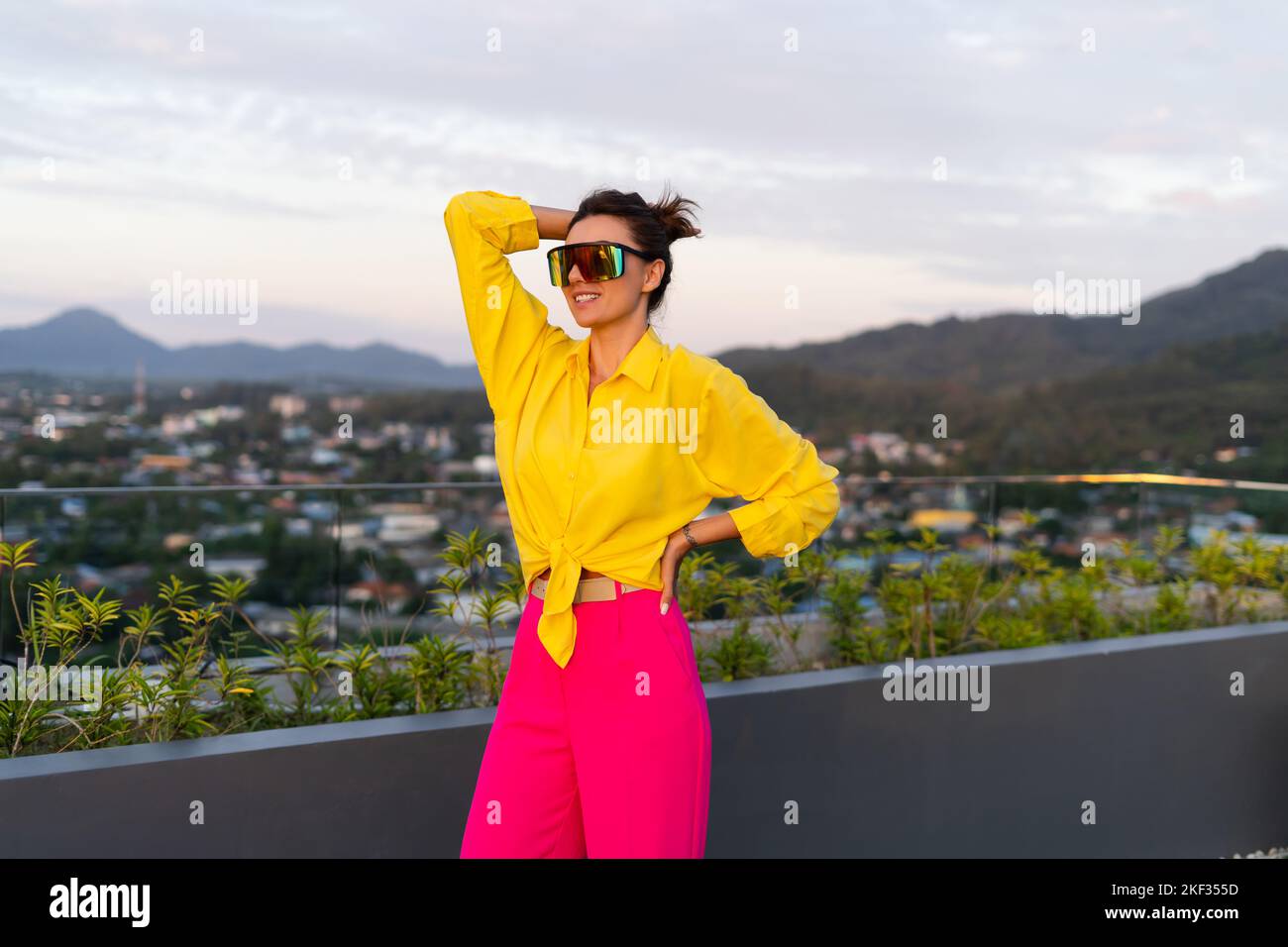 Chic tenue des femmes de mode dans le pantalon de jambe large rose vif et  chemise jaune sac de maintien lunettes de soleil tendance posant sur le  toit terrasse vue tropicale extérieur