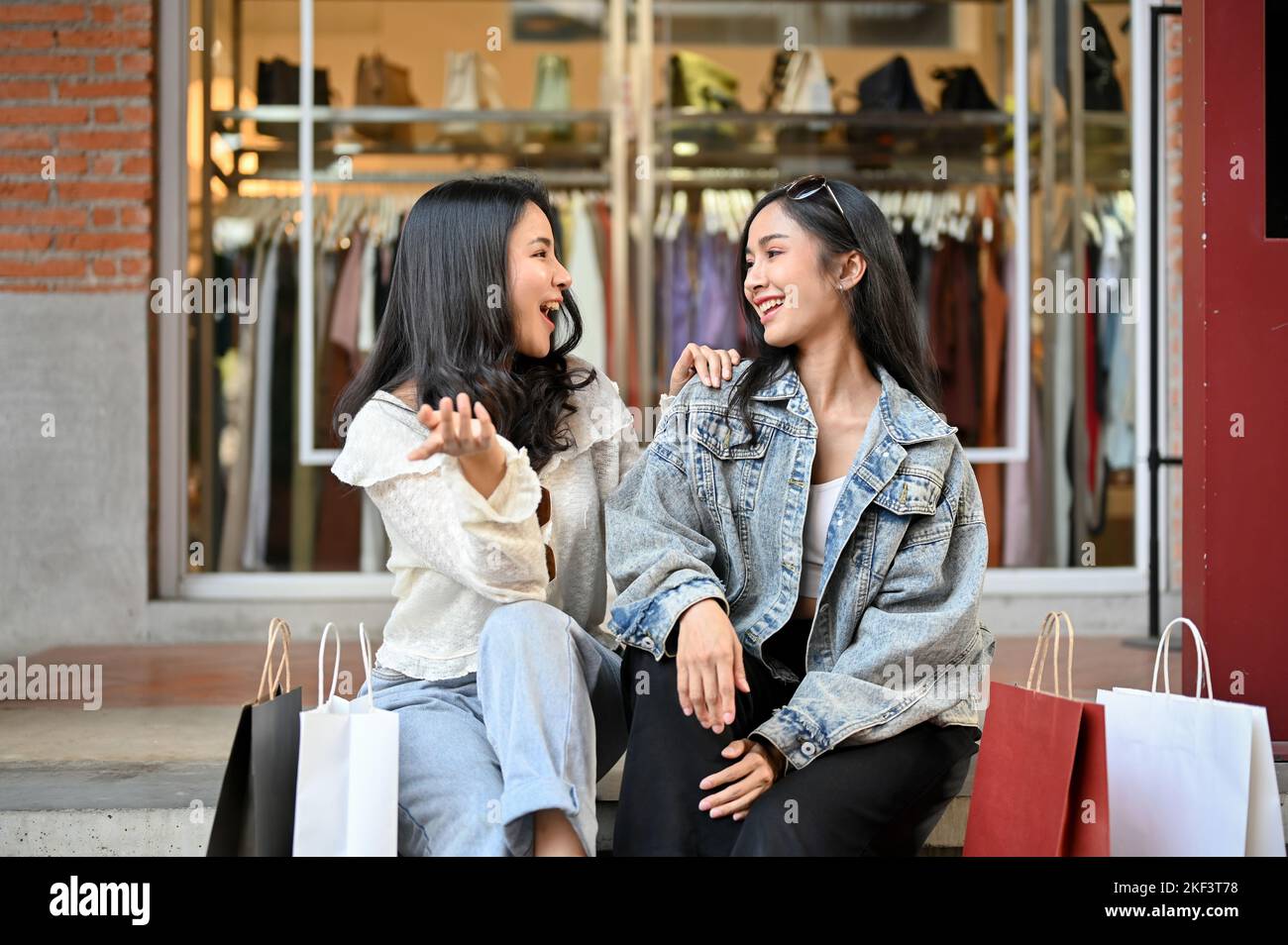 Deux jeunes femmes asiatiques surjoyeuses et heureuses aiment parler et rire tout en s'asseyant sur les escaliers en face du magasin de clothe ensemble Banque D'Images