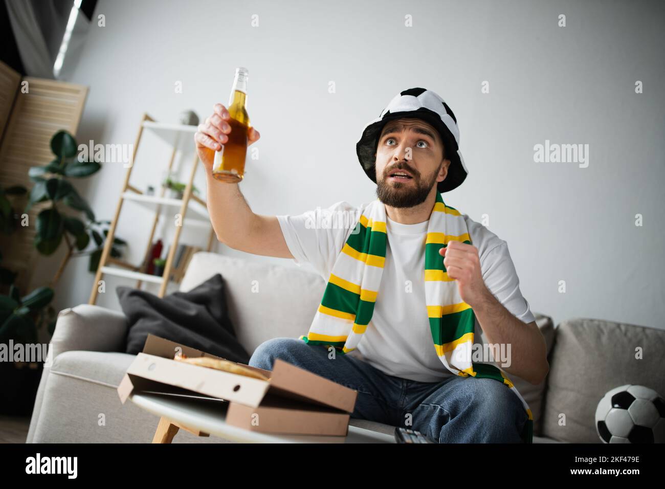 homme tensionné dans un chapeau de fan sportif et un foulard tenant une bouteille de bière tout en regardant le championnat, image de stock Banque D'Images