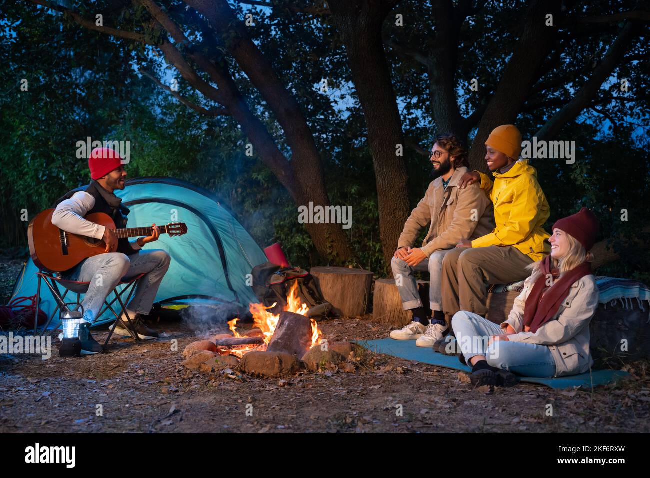 Des amis multiethniques heureux s'amusant à jouer de la musique et à profiter du camping de feu de joie dans la nature la nuit Banque D'Images