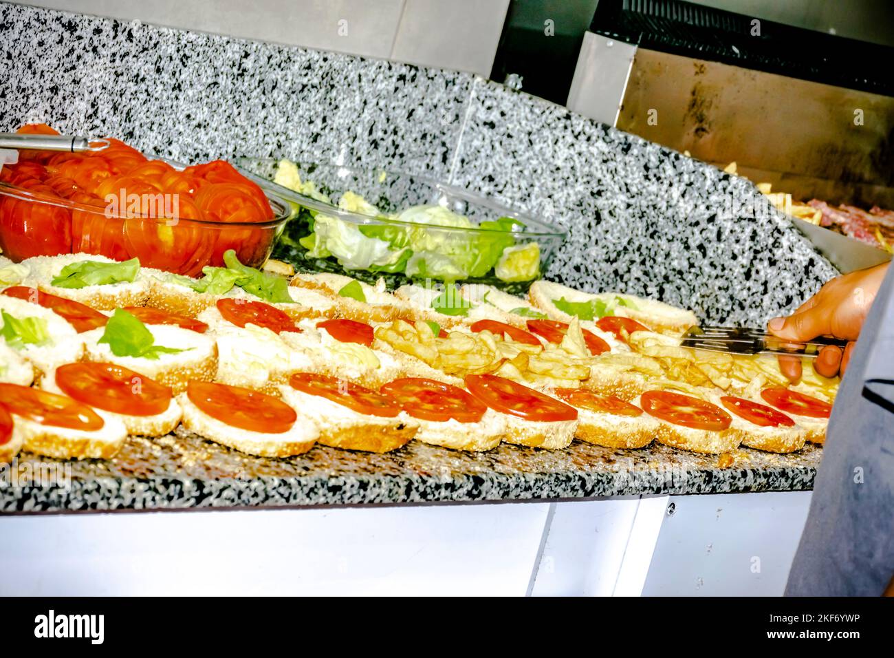 Des assiettes de laitue et de tomate sur le plan de travail, ouvraient les hamburgers d'affilée avec des frites et des tranches de tomate. Cuisinière main tenant une table de cuisine et les remplissant Banque D'Images