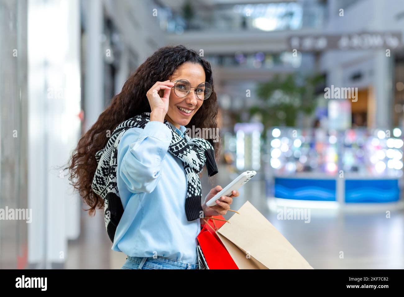 Femme d'affaires magasiner. Jeune femme hispanique marchant et magasiner dans un centre commercial. Il tient un téléphone et des paquets colorés avec des marchandises dans ses mains, tient sur ses lunettes, regarde la caméra, et sourit. Banque D'Images