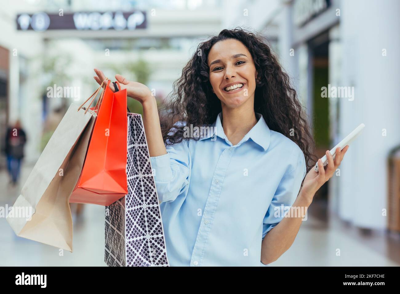 Une jeune femme hispanique attrayante fait ses courses dans un centre commercial, avec des sacs colorés et un téléphone portable. Affiche les achats, les brags, les sourires. Banque D'Images