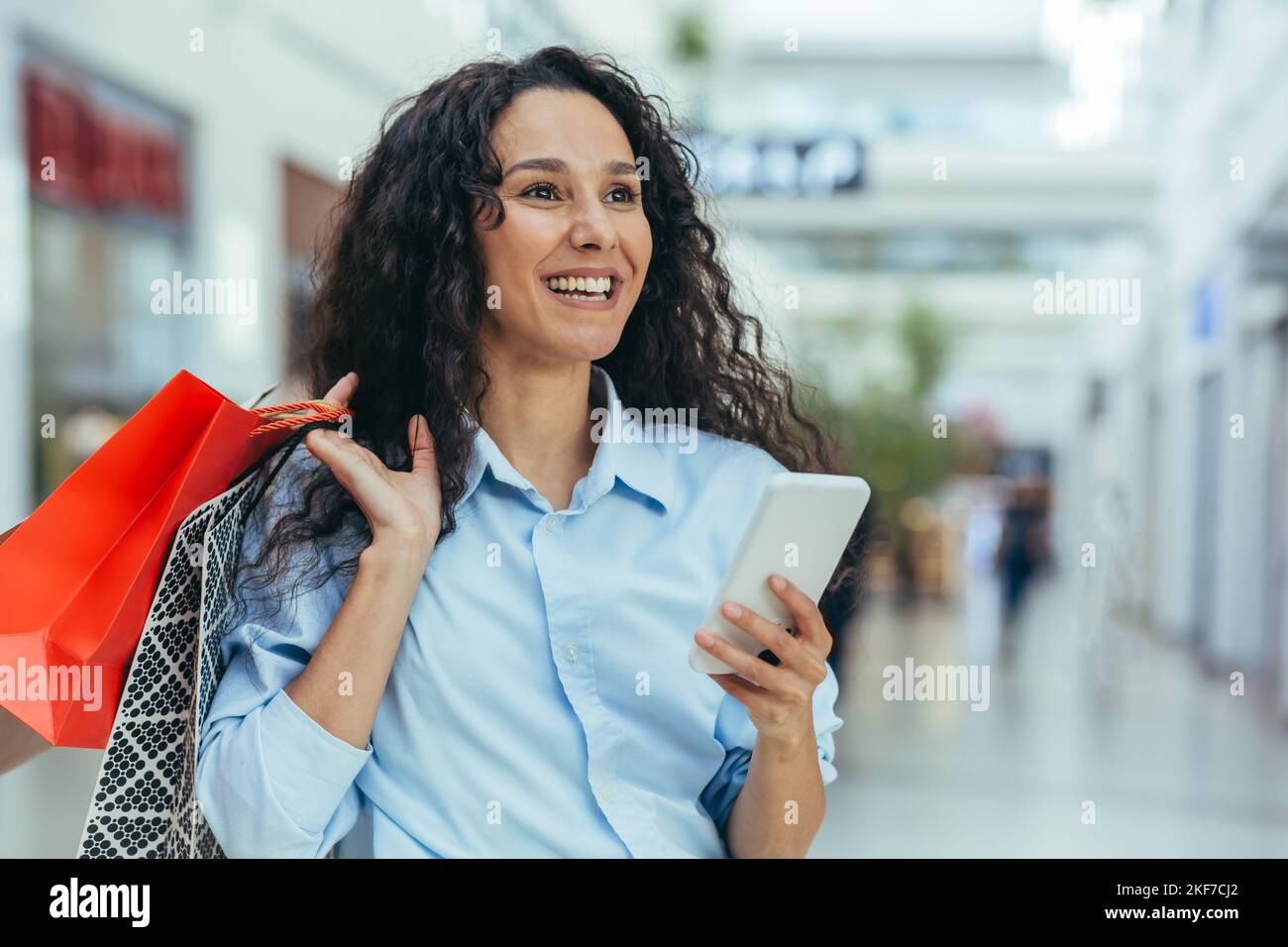 Jeune femme hispanique attrayante et satisfaite magasiner dans un centre commercial, tenant des sacs colorés et un téléphone portable. Appels, types de messages, paiement des achats en ligne. Banque D'Images