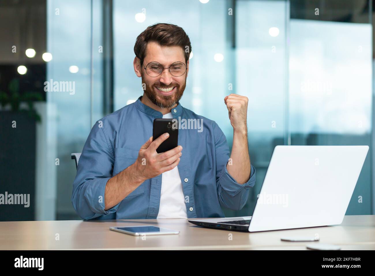 Joyeux jeune homme beau indépendant jouant des jeux en ligne sur le téléphone au travail. Lève la main, montre oui, gagné. Assis dans le bureau au bureau. Banque D'Images