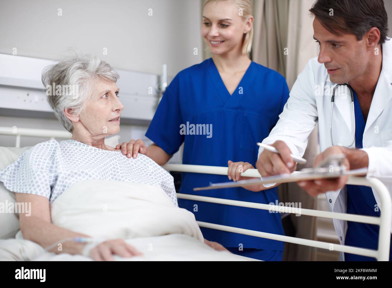 Je suis bien. Un médecin et sa jeune infirmière montrant à leur patient son rapport médical en se tenant debout au-dessus de son lit. Banque D'Images