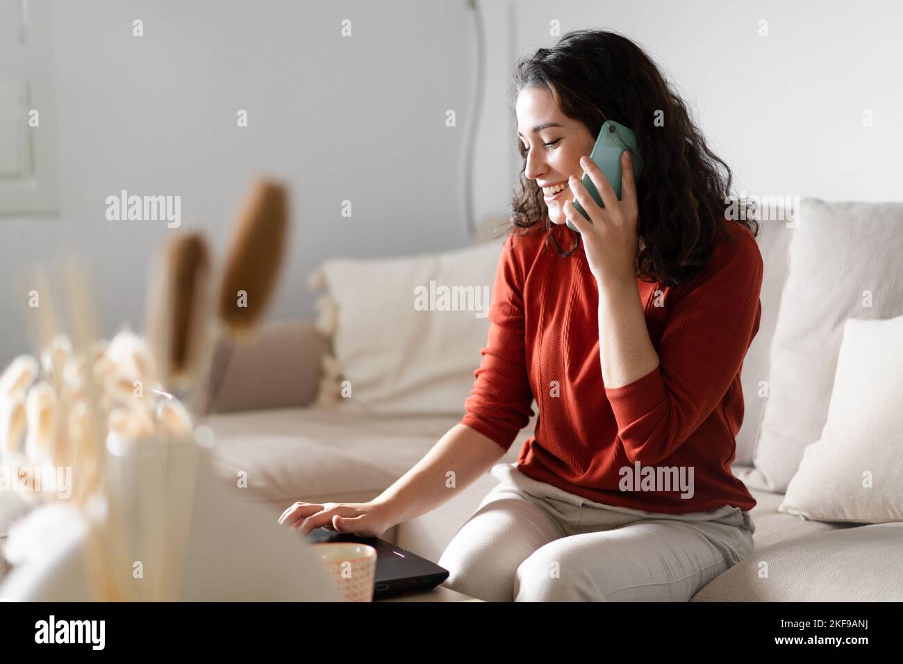 Jeune femme heureuse parlant au téléphone et tapant sur l'ordinateur assis sur le canapé à la maison Banque D'Images