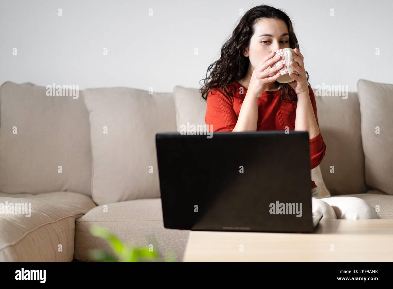 Jeune femme ayant un café et regardant de côté l'écran d'ordinateur assis sur le canapé à la maison. Concept de travail à domicile Banque D'Images