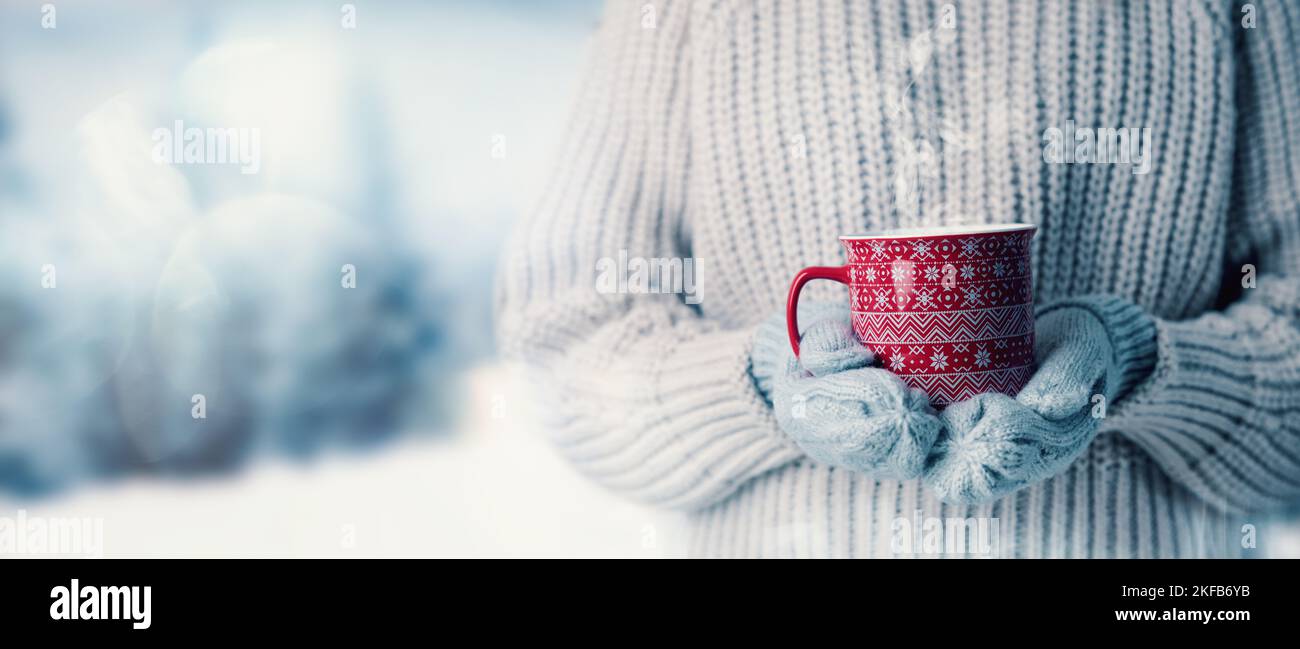femme en laine chandail et gants mitaines tenant une tasse de boisson chaude à la vapeur sur fond de paysage hivernal neigeux. bannière avec espace de copie Banque D'Images