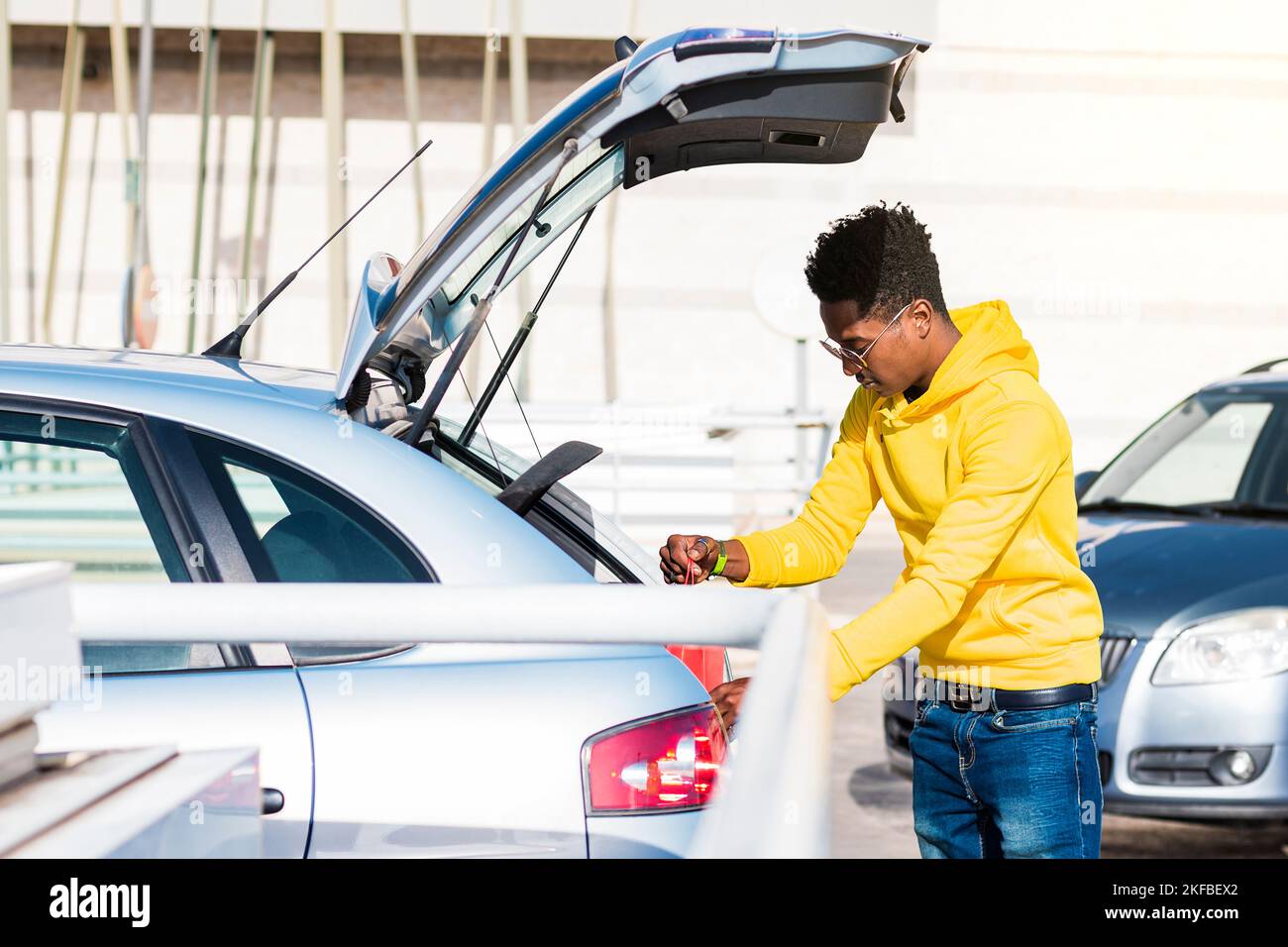 Jeune Africain fumant vêtu d'un sweat-shirt jaune, d'un Jean bleu et de lunettes de soleil portant un sac en papier rouge dans le coffre d'une voiture dans la rue. Banque D'Images