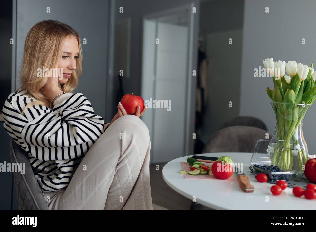 La jeune blonde enceinte dans les vêtements de la maison est assise à table dans sa cuisine et tient la pomme juteuse rouge dans ses mains. Banque D'Images