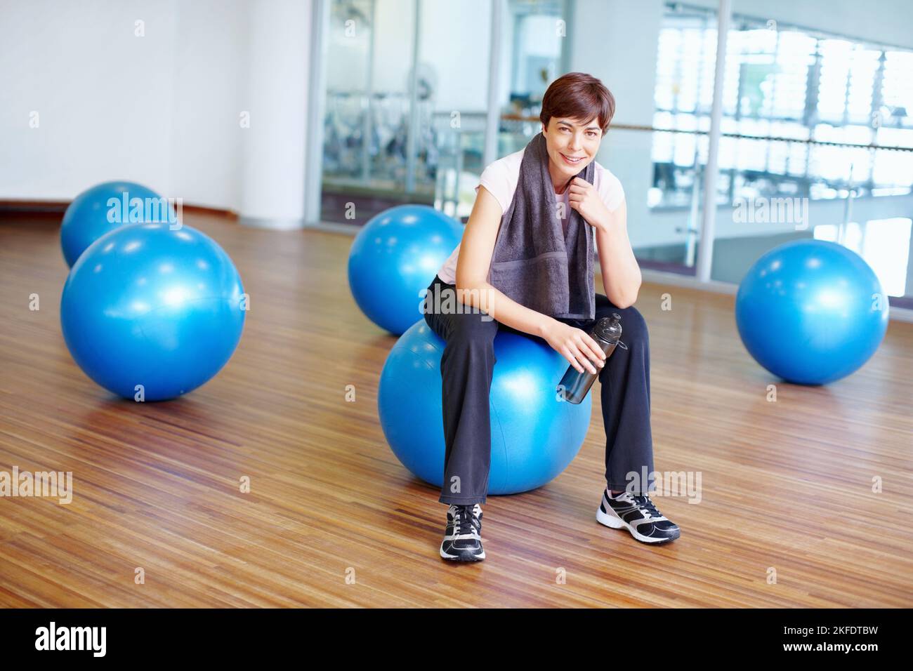 Faire une pause pendant l'entraînement. Portrait d'une femme assise sur un ballon d'exercice avec une serviette et une bouteille d'eau. Banque D'Images