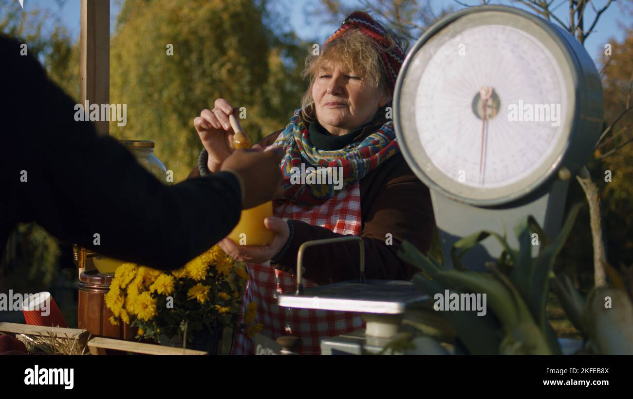 La vendeuse âgée se tient à la stalle, donne le miel au goût. Les gens paient pour un pot de miel avec de l'argent. Marché agricole local ou foire d'automne en plein air. Plats végétariens et bio. Agriculture. Banque D'Images
