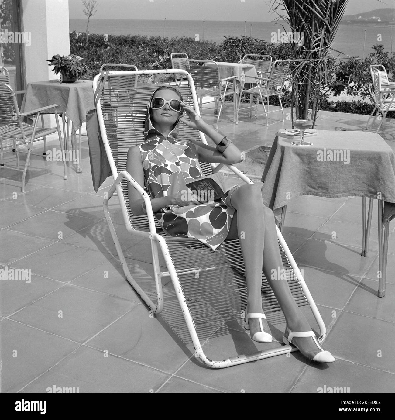 Tournage de mode sur l'île espagnole Ibiza en 1960s. Un modèle de mode sur place modélise la robe d'été typique, en profitant du soleil assis sur la terrasse de l'hôtel. Elle porte les grandes lunettes de soleil tendance de la décennie. 1967 Banque D'Images