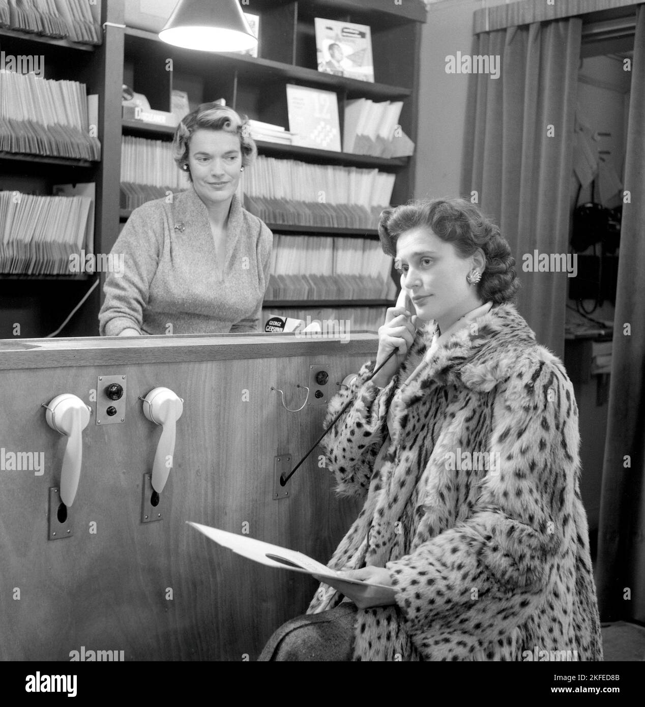Comment vous avez acheté des disques dans le 1950s. La dame tenant ce qui ressemble à un téléphone écoute en fait un enregistrement. À ce moment-là, il était courant que les clients puissent écouter les dossiers avant de décider des acheter. Le magasin de disques dispose de plusieurs stations d'écoute et d'une femme derrière le comptoir gérant le système. Elle détient un disque portant le nom de George Gershwin. Suède 1950s. Banque D'Images