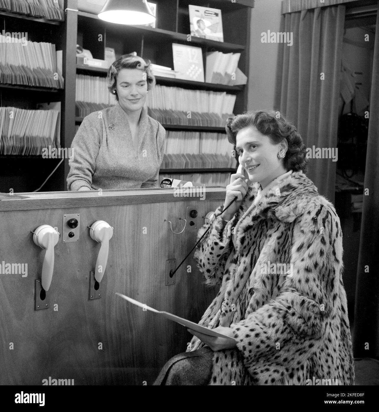 Comment vous avez acheté des disques dans le 1950s. La dame tenant ce qui ressemble à un téléphone écoute en fait un enregistrement. À ce moment-là, il était courant que les clients puissent écouter les dossiers avant de décider des acheter. Le magasin de disques dispose de plusieurs stations d'écoute et d'une femme derrière le comptoir gérant le système. Elle détient un disque portant le nom de George Gershwin. Suède 1950s. Banque D'Images