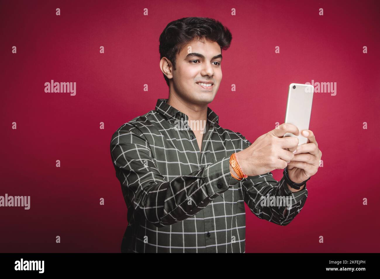 Un jeune Indien souriant dans une chemise à carreaux prend un selfie sur un smartphone sur un fond rouge. Lors d'un appel vidéo avec une petite amie ou une fiancée. Banque D'Images