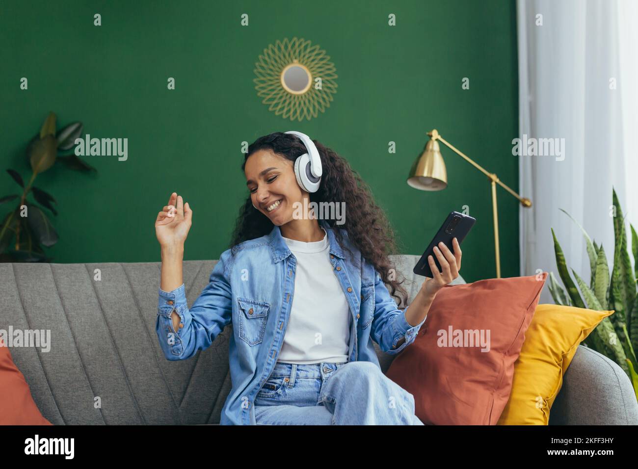 Une jeune femme attirante d'Amérique latine écoute de la musique dans des écouteurs blancs, s'assoit sur le canapé, danse, tient le téléphone dans ses mains. Banque D'Images