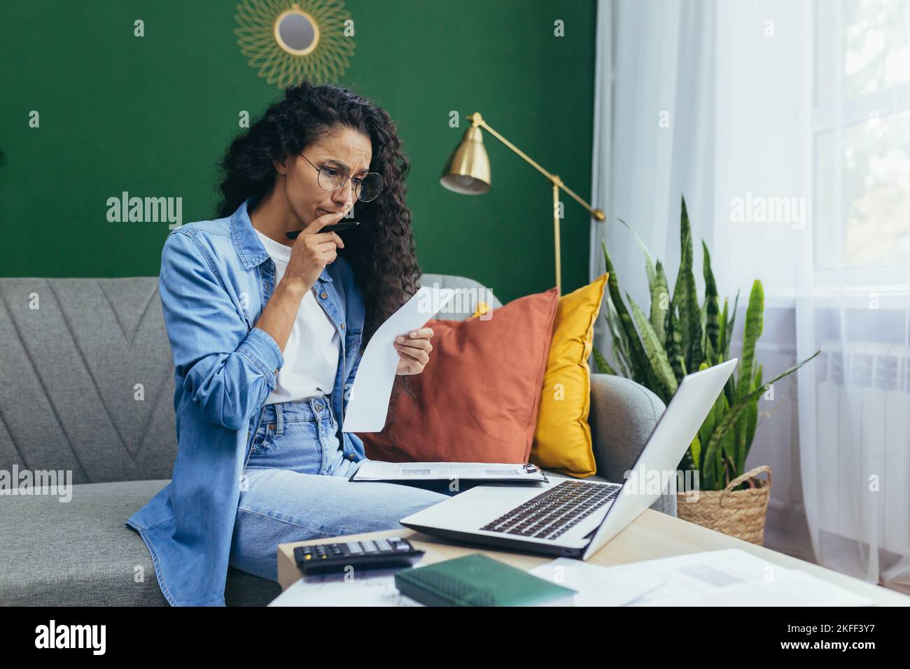 Jeune femme d'affaires hispanique concentrée et sérieuse, indépendante travaillant à la maison sur ordinateur portable, tenant des documents en mains, assis sur un canapé. Banque D'Images