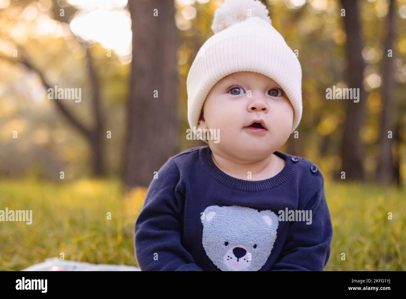 Bébé en bonnet blanc tricoté est assis sur l'herbe dans le parc contre le fond des arbres d'automne Banque D'Images