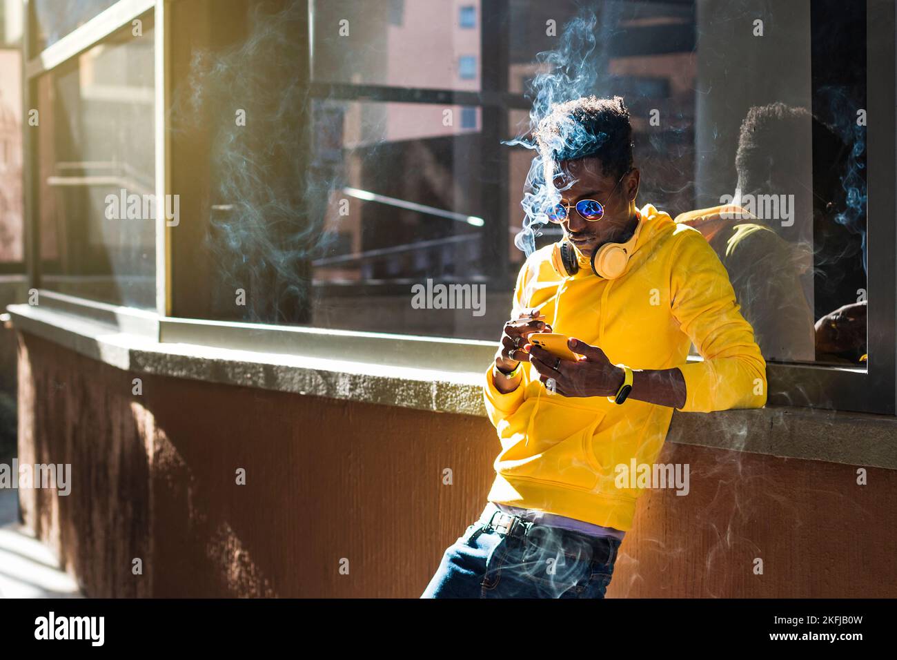 Un jeune Africain vêtu d'un sweat-shirt jaune, d'un Jean bleu et de lunettes de soleil se penche sur un rebord de fenêtre regardant son téléphone portable tout en tenant un c Banque D'Images