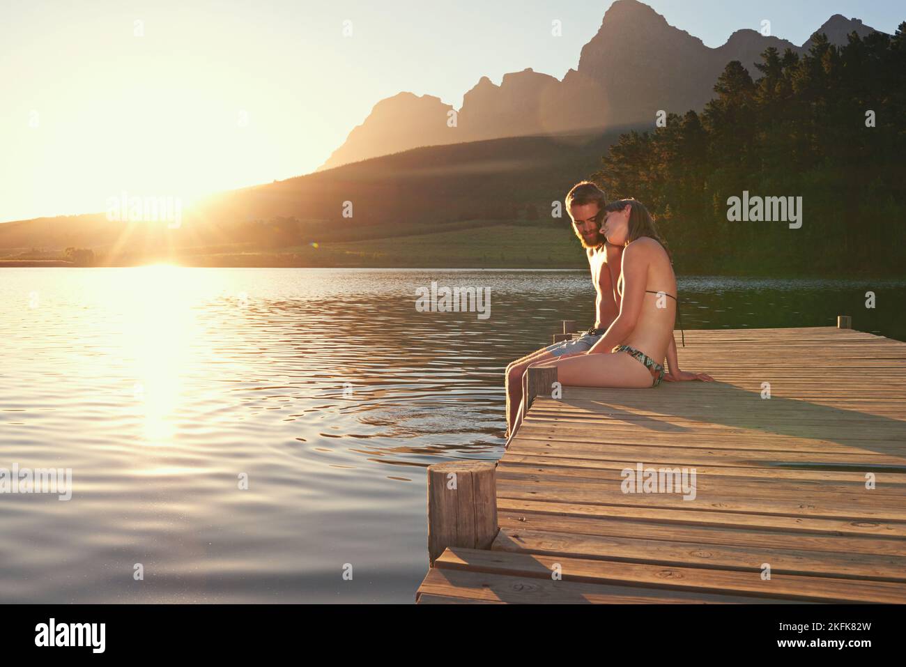 Amour au bord du lac. Jeune couple affectueux en maillots de bain assis sur un quai au coucher du soleil. Banque D'Images