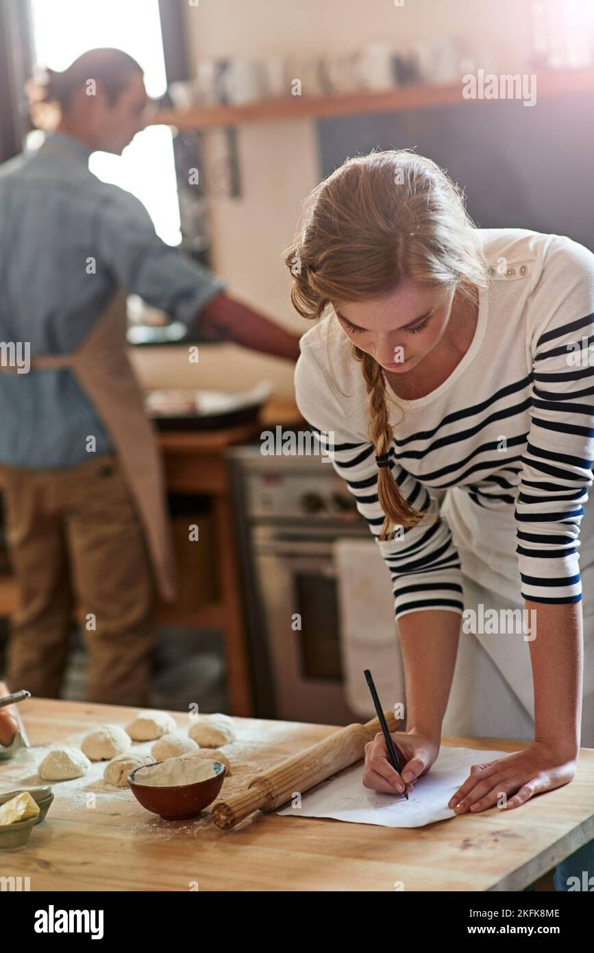 L'ingrédient secret est une cuillère à café d'amour. Une femme écrivant une recette pour la pâte qui se prépare avec son petit ami en arrière-plan. Banque D'Images