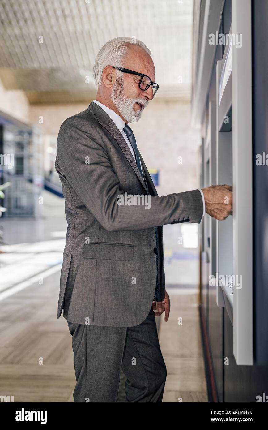 Vue latérale d'un homme âgé professionnel utilisant un distributeur automatique de billets. Homme d'affaires senior portant des lunettes tout en retirant de l'argent liquide à un distributeur automatique de billets. Il porte des Banque D'Images