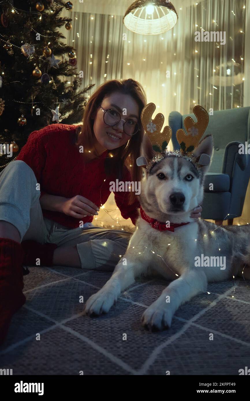 Jeune femme le soir de Noël avec chien d'animal de compagnie ayant le plaisir de mettre sur les cornes de cerf de Noël et guirlande assis sur le sol à Noël Banque D'Images