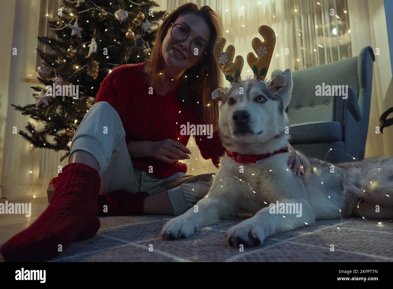 Jeune femme le soir de Noël avec chien d'animal de compagnie ayant le plaisir de mettre sur les cornes de cerf de Noël et guirlande assis sur le sol à Noël Banque D'Images