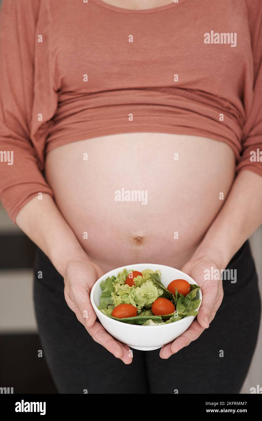 Juste pour le bébé. Une jeune femme enceinte tenant une salade dans la cuisine. Banque D'Images