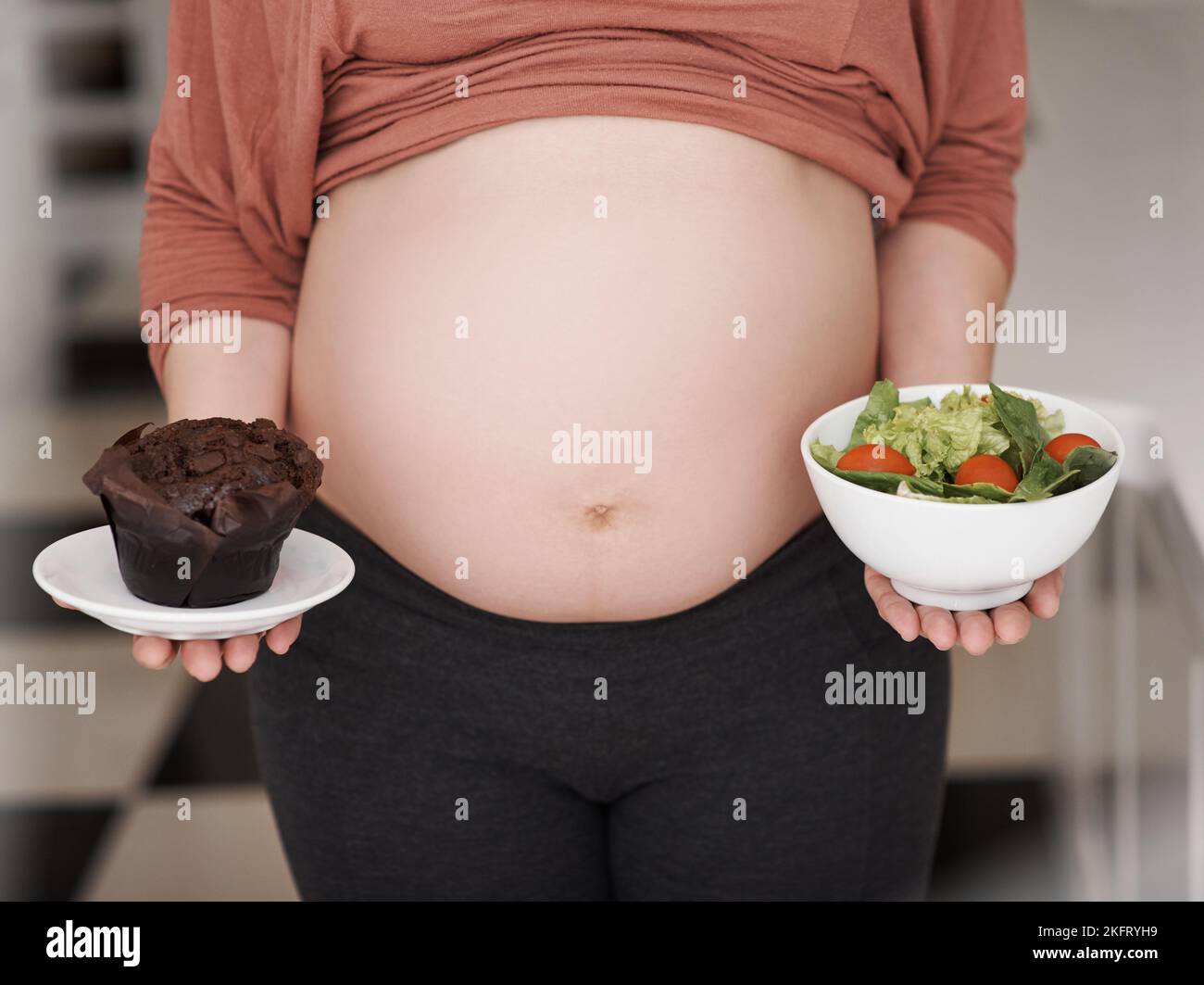 Faites le bon choix. Une jeune femme enceinte qui décide entre une salade ou un cupcake dans la cuisine. Banque D'Images