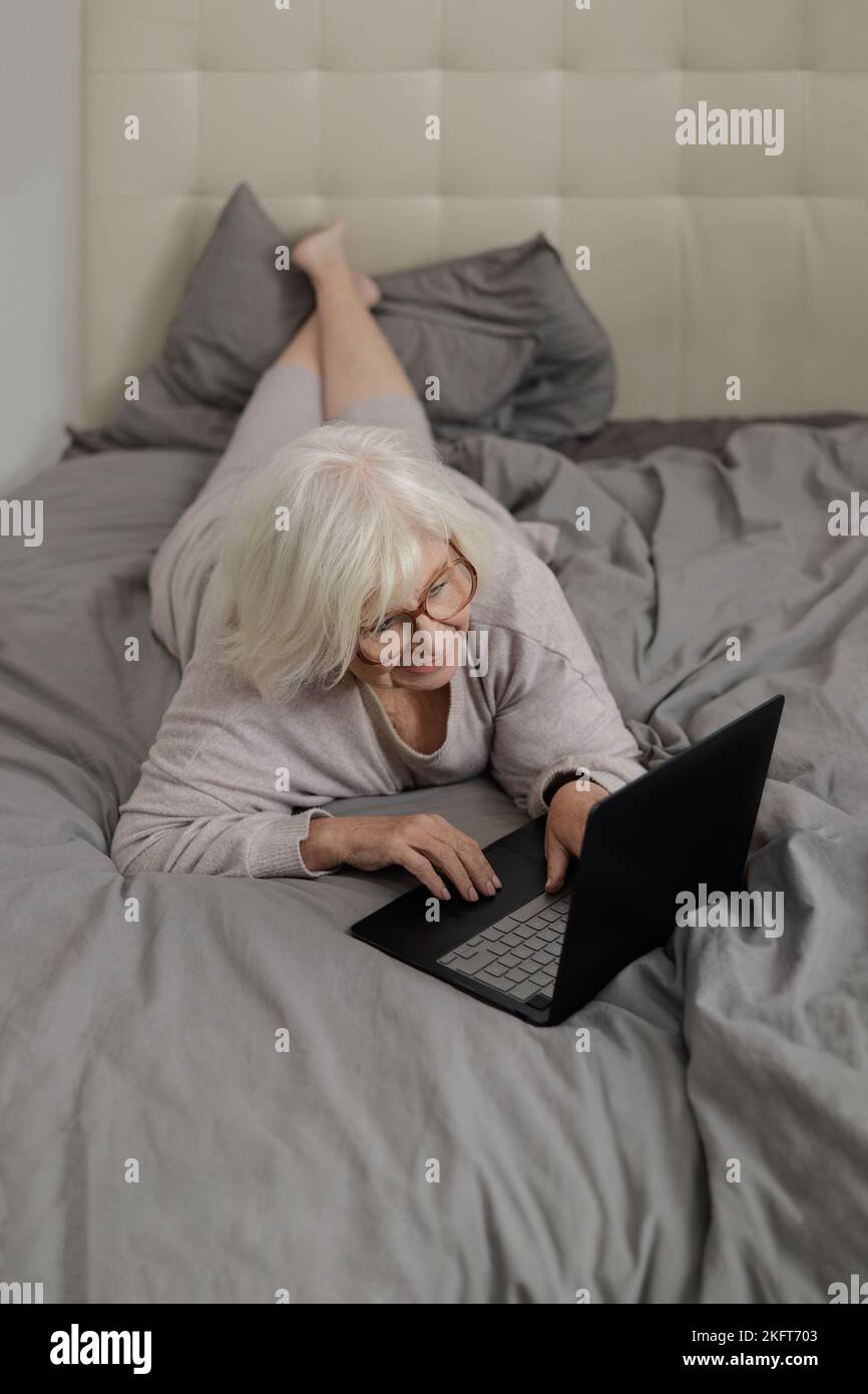 De dessus tout le corps de la femme âgée concentrée dans les lunettes couché sur le lit mou avec des vêtements de lit gris et de taper des informations sur ordinateur portable à la maison Banque D'Images