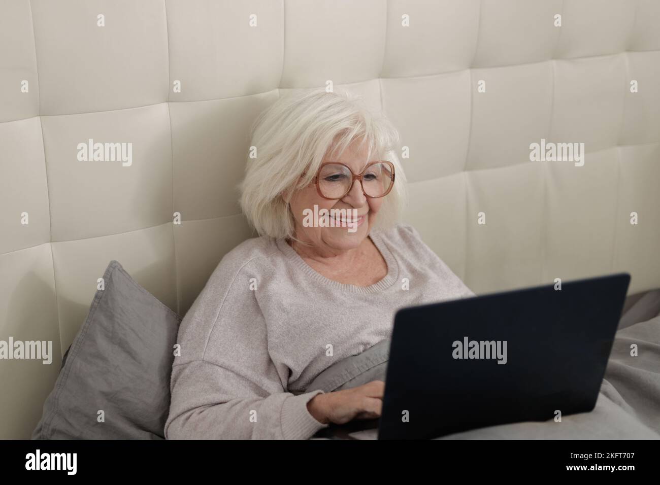Bonne dame âgée en lunettes de vue couchée sur un lit souple avec des couvre-lits gris et des informations dactylographiées sur un ordinateur portable à la maison Banque D'Images