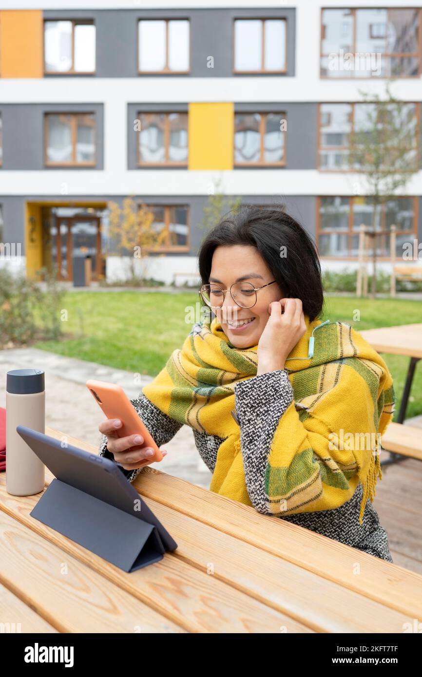 Jeune femme utilisant des écouteurs et un téléphone portable assis à table avec une tablette et un en-cas pour déjeuner dans le parc du campus en automne Banque D'Images