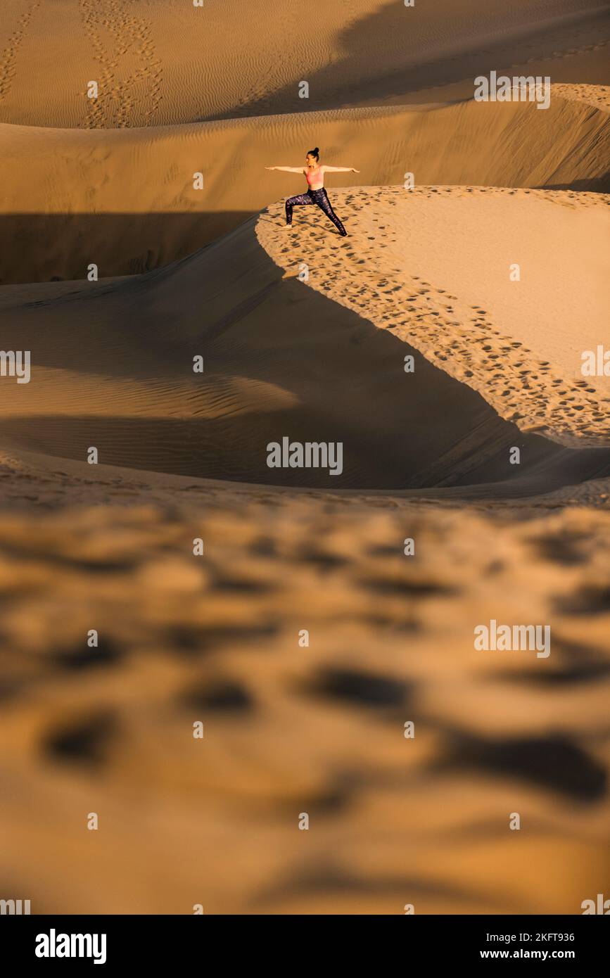 Vue de loin de la jeune femme pratiquant le yoga sur les dunes de sable dans le désert. Posture du guerrier. Banque D'Images