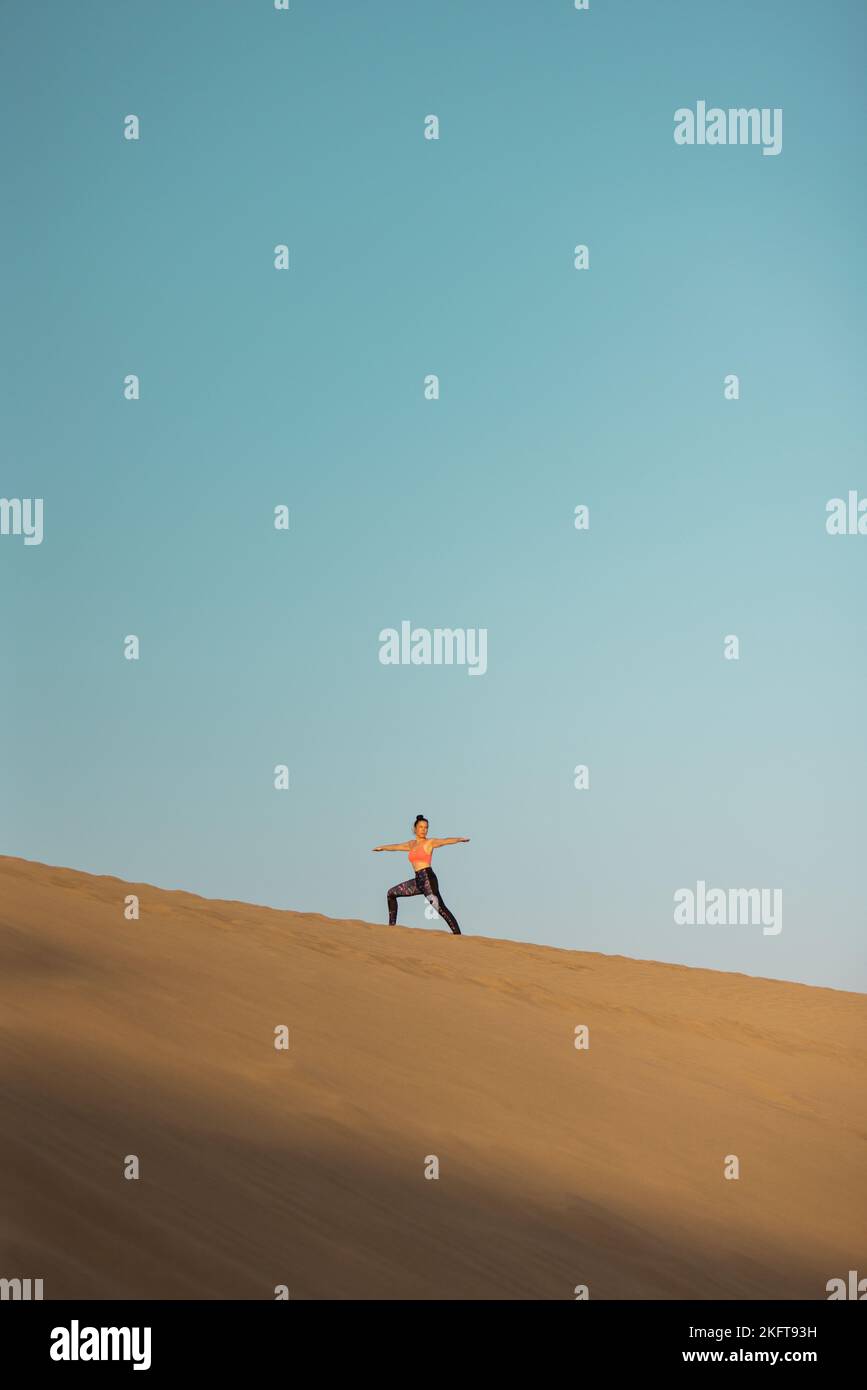 Vue de loin de la jeune femme pratiquant le yoga sur les dunes de sable dans le désert. Posture du guerrier. Banque D'Images