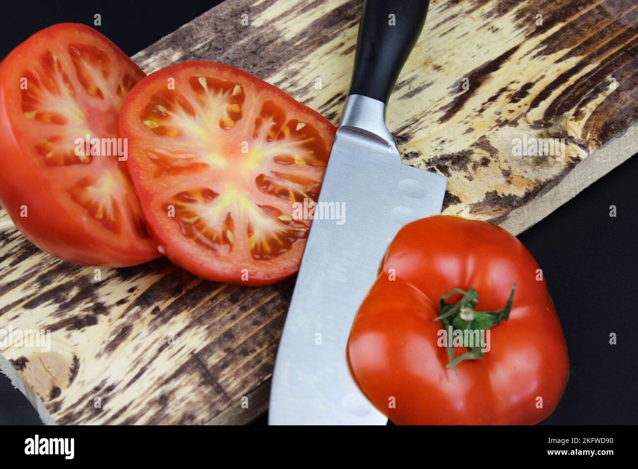 Vue en grand angle des tranches de tomates avec couteau sur le plan de coupe sur fond sombre. Planche à découper en bois avec tomates de légumes crus sur Boa à découper rustique Banque D'Images