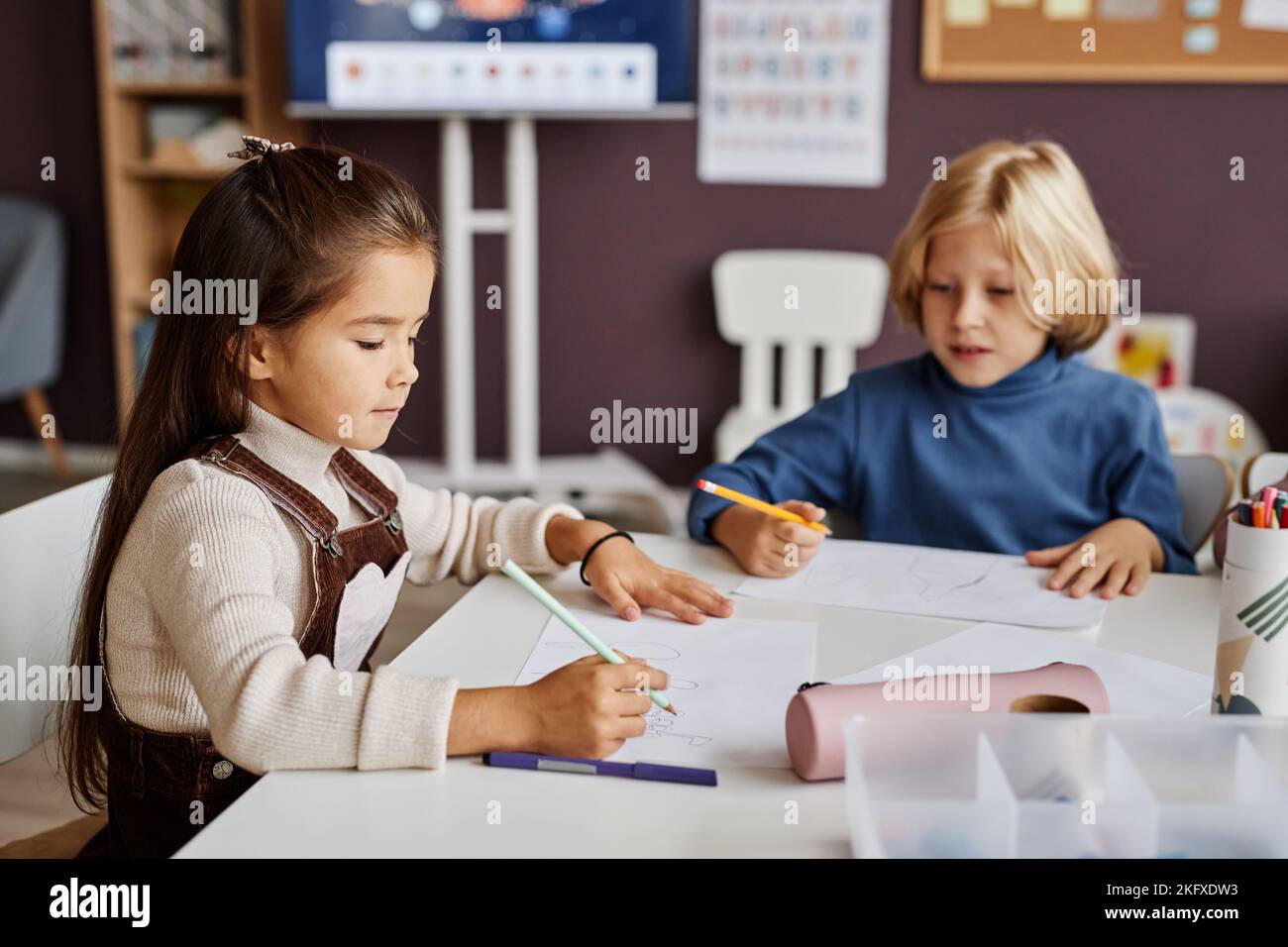 Deux écoliers diligents qui dessinant sur des papiers avec des crayons tout en étant assis à table dans la salle de classe de la maternelle ou maternelle moderne Banque D'Images
