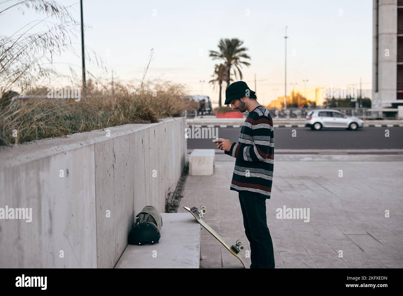 un homme caucasien avec piercing de nez en chandail rayé regardant son écran de smartphone dans la ville à côté de son skateboard Banque D'Images