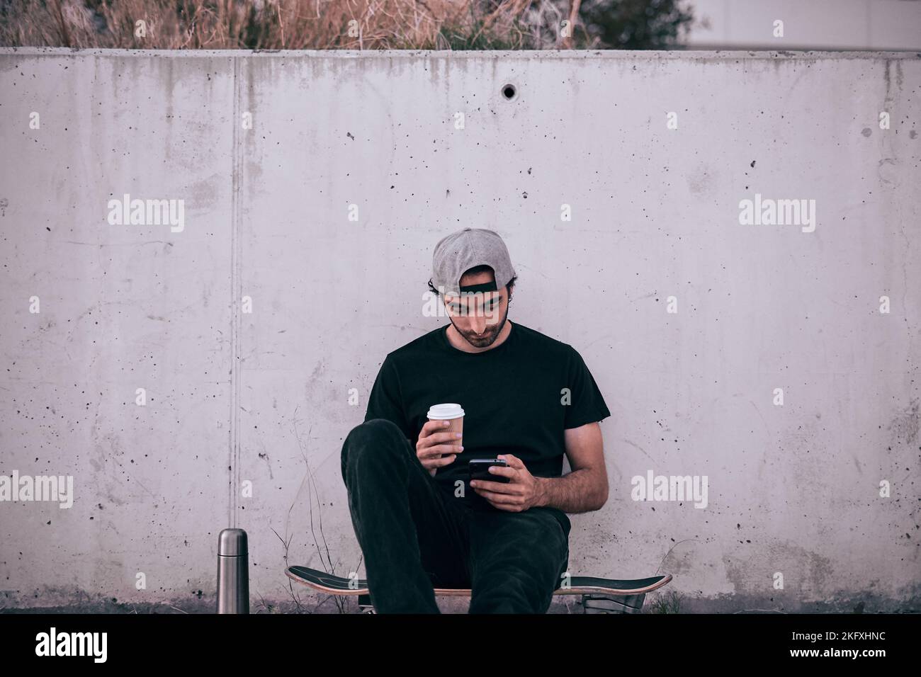 homme caucasien en casquette grise et t-shirt noir assis sur son skateboard manipulant son smartphone avec sa main gauche et tenant un verre de café Banque D'Images