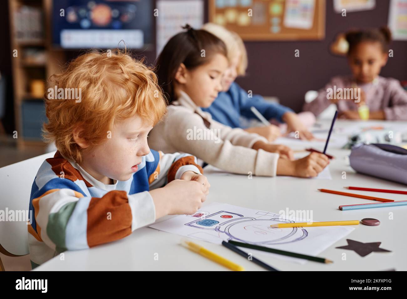 Rangée d'apprenants interculturels diligents de l'école maternelle assis par table et en dessinant avec des crayons avec mignon petit garçon à poil de gingembre devant Banque D'Images