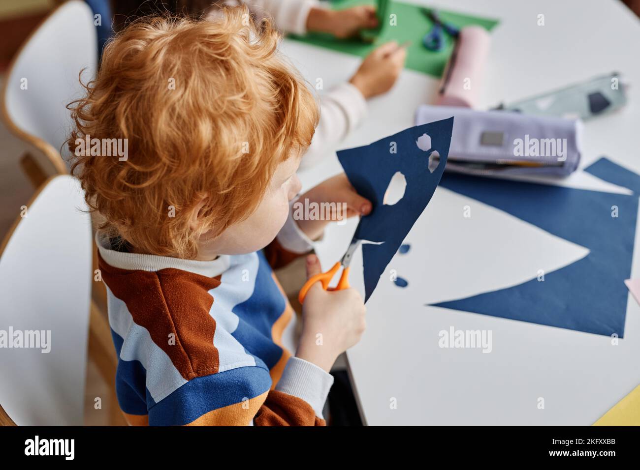 Au-dessus de l'angle de cute diligent école de coupe des trous dans le papier bleu foncé tout en créant le masque à la leçon dans l'école maternelle ou maternelle Banque D'Images